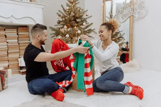 Couple exchanging Christmas sweaters in festive holiday setting with tree.