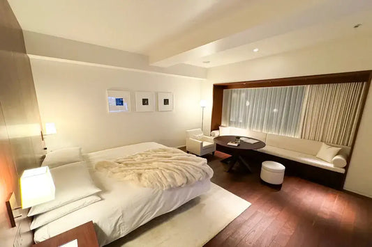 Modern hotel room with king-sized bed, plush beige blanket, cozy seating area, and warm lighting.