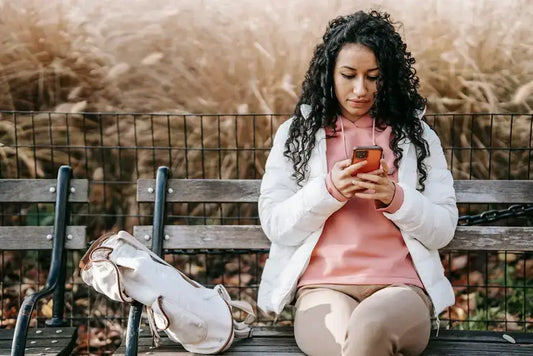 Woman in pink hoodie using smartphone on park bench with backpack nearby