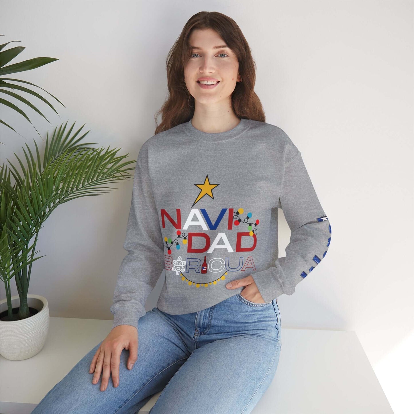 Woman wearing a grey Christmas sweater with "NAVIDAD BORICUA" design and festive prints on left sleeve, sitting by a plant.