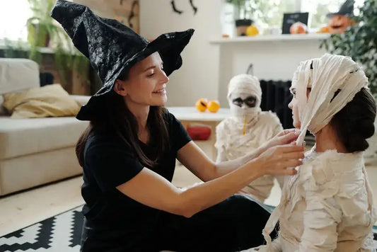 Woman in witch hat helping child dress as mummy for Halloween in cozy living room.