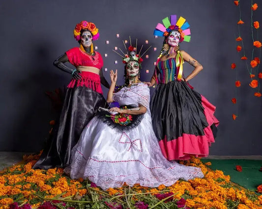 Three women in vibrant traditional Mexican costumes with colorful patterns and makeup, surrounded by marigold flowers.