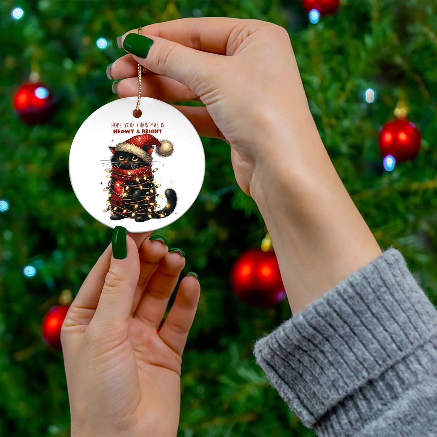 Person holding a circular Meowy Xmas ceramic ornament featuring a cat, with a Christmas tree in the background.