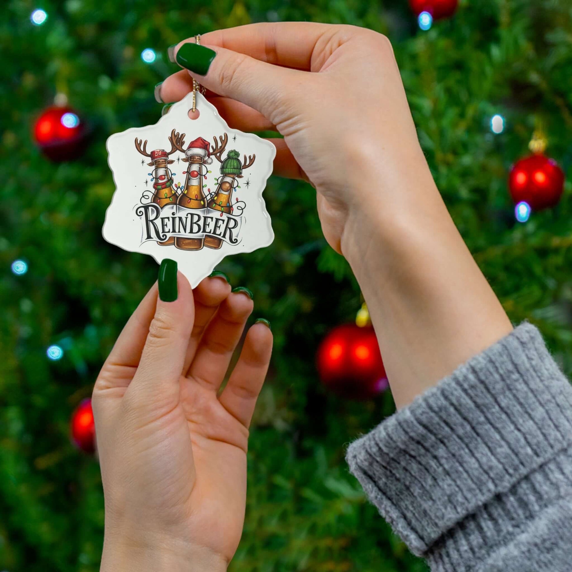 Hands holding a vibrant Reinbeer ceramic ornament, featuring festive reindeer, against a Christmas tree backdrop with ornaments.