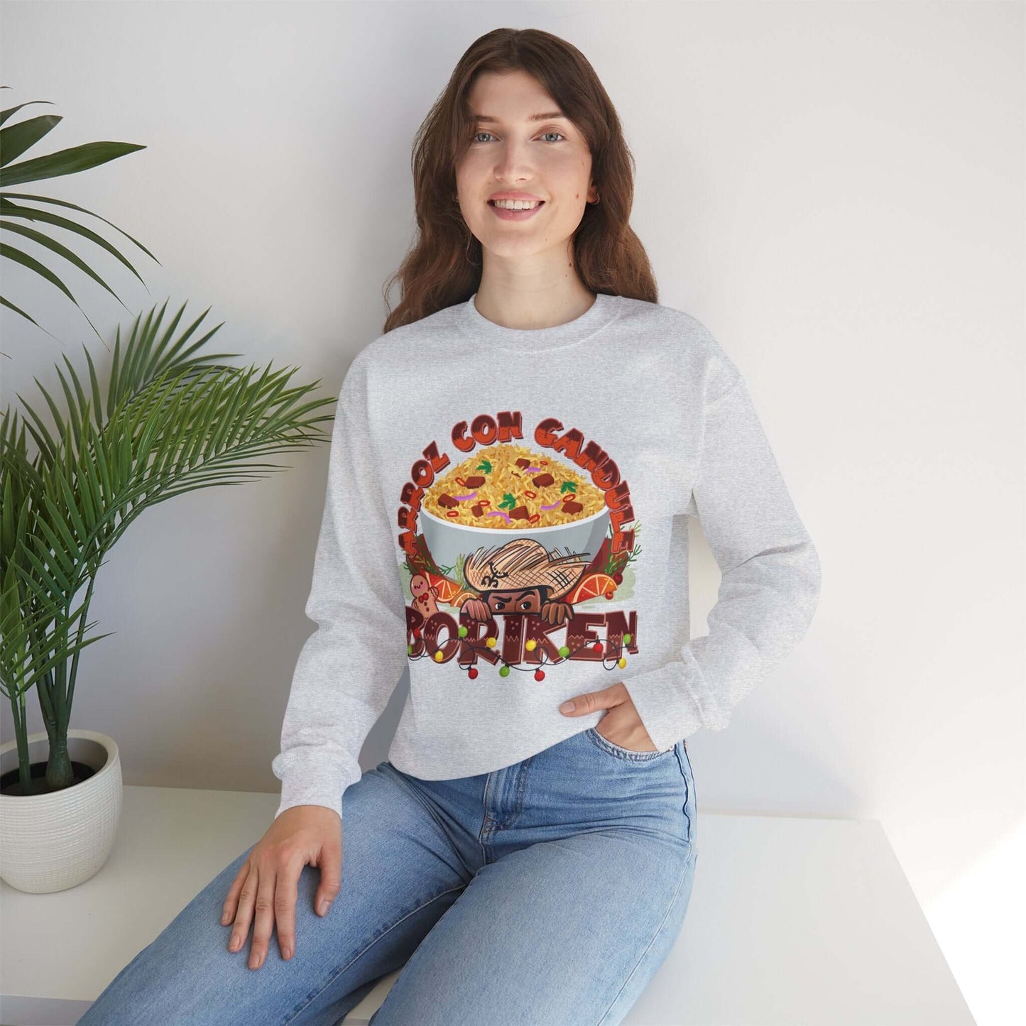 Woman wearing Arroz Con Gandules Christmas Sweater with festive rice bowl graphic and Borikén text, sitting in a bright room.